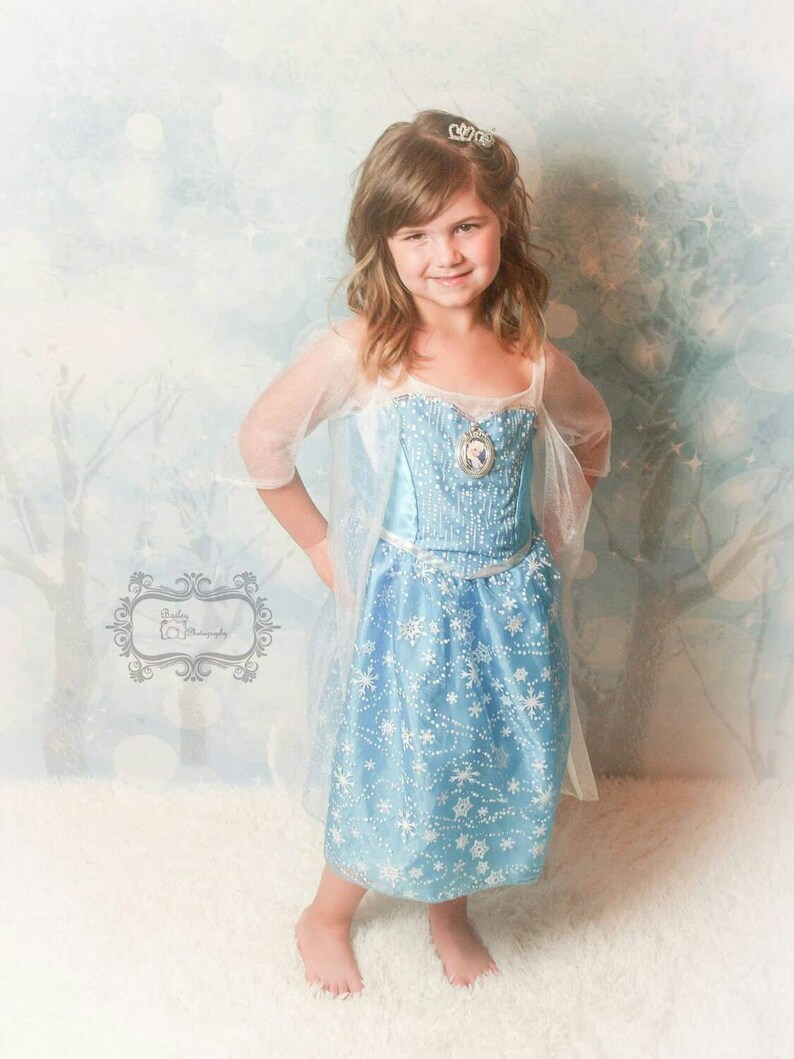 May include: A young girl wearing a blue and white snowflake patterned dress with a sheer overlay and a silver tiara. She is standing on a white fluffy surface.