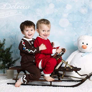 May include: Two young boys in holiday pajamas are riding a vintage wooden sled. The boy in the red pajamas is steering the sled. The boy in the brown pajamas is wearing a gingerbread man costume. A white plush snowman is in the background.