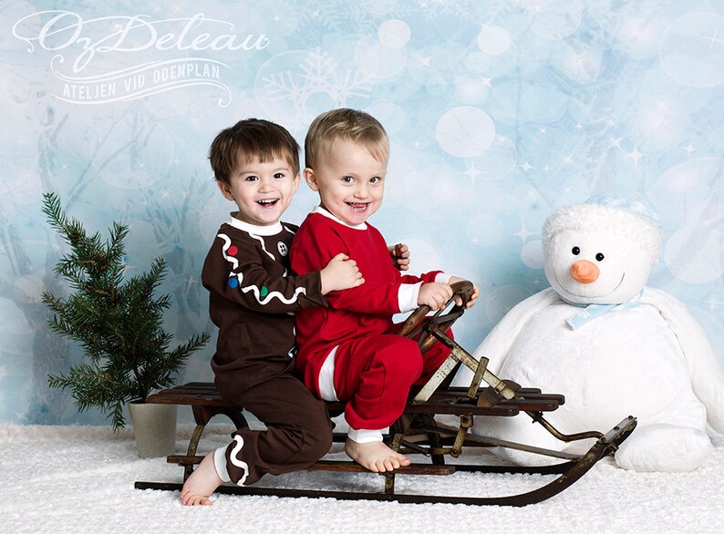 May include: Two young boys in holiday pajamas are riding a vintage wooden sled. The boy in the red pajamas is steering the sled. The boy in the brown pajamas is wearing a gingerbread man costume. A white plush snowman is in the background.