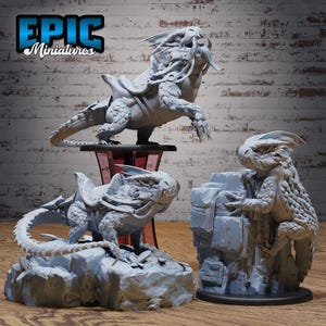 May include: Three gray miniature dragon figurines. One dragon is mounted on a saddle, another is perched on a rock, and the third is climbing a rock formation. The text "EPIC Miniatures" is in the upper left corner.