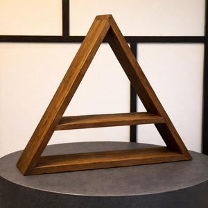 May include: A dark brown wooden triangular shelf unit with two internal shelves. The geometric design is set against a white and black backdrop. The shelf is designed for wall mounting and is made of wood.