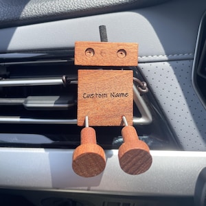Könnte beinhalten: Ein holzförmiger Roboter-Auto-Lufterfrischer. Der Roboter hat einen rechteckigen Körper mit dem eingravierten Text "Custom Name". Er hat einen rechteckigen Kopf mit zwei kreisförmigen Augen. Die Beine des Roboters sind zylindrisch.