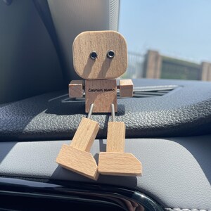 May include: A wooden robot figurine with a square head, arms, and legs. The eyes are represented by two dark circles. The text "Custom Name" is engraved on the chest. The legs are connected to the body with metal rods. The figurine is sitting on a car dashboard.