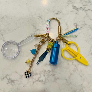 May include: A gold-toned metal keychain featuring a variety of charms including a clear magnifying glass, a blue cylindrical pill container, yellow plastic scissors, a blue seam ripper, and a heart charm with a black and white checkerboard pattern.