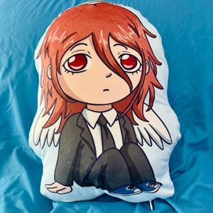 May include: A plush pillow in the shape of an anime character with red hair and large red eyes. The character is wearing a black suit, white shirt, and blue sneakers. White angel wings are visible.