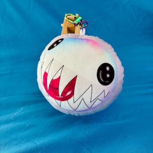 Puede incluir: Un llavero de peluche blanco y redondo con una cara de dibujos animados. Tiene ojos negros, una boca roja con dientes afilados y un degradado arcoíris. Lleva un cierre de arcoíris.