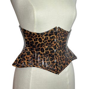 May include: A leopard print corset with a glossy finish. The corset features a structured design with a pointed hemline and is fastened with small, silver-toned buttons down the front. The garment is displayed on a mannequin.