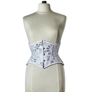 May include: White corset with silver floral embroidery, displayed on a mannequin. The corset features a structured design with a pointed waistline and a series of small buttons down the front. The fabric has a subtle sheen, and the overall aesthetic is elegant.