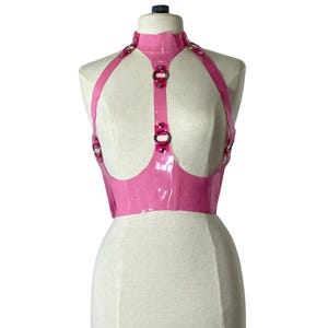 Puede incluir: Un arnés de látex rosa brillante con cuello alto y correas. El arnés presenta anillos de metal plateado y se muestra en un maniquí. La prenda está diseñada para ser usada como top.