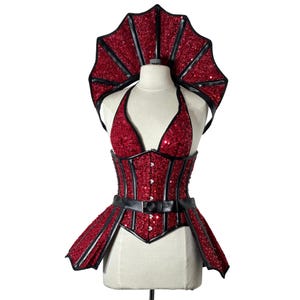 Op de afbeelding: Een rood paillettenkorset en bijpassende hoofdtooi met zwarte rand. Het korset heeft een haltertop en een zwarte riem. De hoofdtooi heeft een waaier vorm. De outfit wordt op een mannequin gepresenteerd.