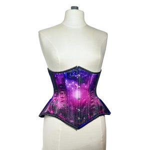 Op de afbeelding: Een paars en roze corset met een galaxy print, getoond op een mannequin. Het corset heeft een zwarte rand en zilverkleurige knopen aan de voorkant. Het ontwerp heeft een levendige mix van kleuren, wat een hemelse esthetiek creëert.