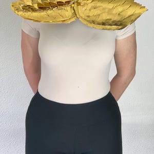 Puede incluir: Un accesorio de cuello dorado con un diseño de capas similar a escamas. La pieza está construida con segmentos reflectantes de acabado metálico que se superponen para crear una apariencia texturizada y alada alrededor del cuello.