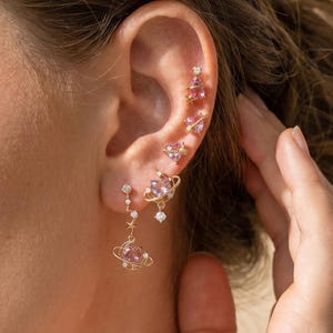 May include: A set of gold-toned earrings featuring pink heart-shaped stones and small clear crystals. The designs include planet-like motifs with rings, star accents, and dangling elements arranged along the earlobe and cartilage piercings.