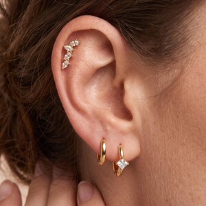 May include: A gold ear climber earring featuring a row of four marquise-cut clear stones worn in the upper cartilage, paired with two gold hoop earrings in the lobe, one of which features a single square-cut clear stone set in the center.