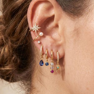 May include: A gold ear stack featuring a starburst ear cuff with clear stones, a small pearl stud, a tiny clear stone stud, and four gold hoop earrings with charms including a blue teardrop, a blue circle, three vertical stones, and a horn.