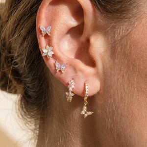 May include: A set of gold-toned earrings featuring two small butterfly studs with iridescent stones, a flower-shaped stud with clear crystals, a small floral-inspired stud, and a hoop earring with a dangling butterfly charm encrusted with clear stones.