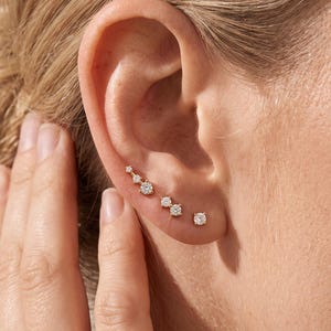 May include: A set of gold-tone earrings featuring a curved climber design with three small clear stones, a two-stone stud, and a single-stone stud, all arranged along the lower edge of the earlobe and cartilage area with a polished finish.