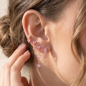 May include: A set of gold-toned earrings featuring pink heart-shaped stones. The collection includes a small stud with a four-pointed star, a single heart drop with a small white bead, a triple heart drop chain, and a small hoop with pink hearts.