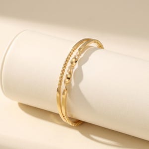 May include: A gold-toned bangle bracelet featuring a dual-strand design. One strand consists of a thin, twisted rope-like texture, while the second strand displays a smooth, undulating wave pattern. The two strands are joined together.