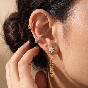May include: A gold-toned ear cuff with a textured surface, a small gold hoop with a single clear stone, and a gold stud earring featuring a six-petaled flower with clear stones and a delicate gold chain hanging from the center of the flower.