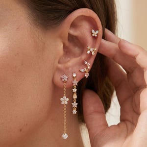May include: A collection of gold-toned earrings worn in a single ear, featuring a butterfly stud, a bow stud, a leaf-shaped climber with a pink heart, a dangling flower chain, and a vertical drop earring with alternating star and round stones.