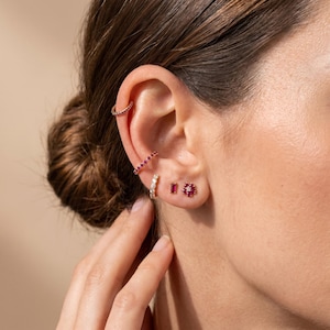 May include: A collection of gold-toned jewelry worn on the ear, including a plain thin ear cuff, a ruby-encrusted ear cuff, a small hoop earring, a rectangular ruby stud, and a flower-shaped stud earring featuring six small rubies and a center stone.