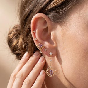 May include: A set of gold-toned earrings featuring celestial motifs, including small star studs, a heart-shaped planet stud with a purple stone, and a dangling crescent moon earring with a purple teardrop stone and orbiting ring detail.
