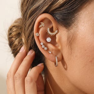 May include: Close-up of an ear adorned with multiple gold and diamond earrings. The ear features a leaf stud, a flower stud, a cluster of three flower studs, a small hoop, a marquise stud, a large round diamond stud, and a gold hoop earring.