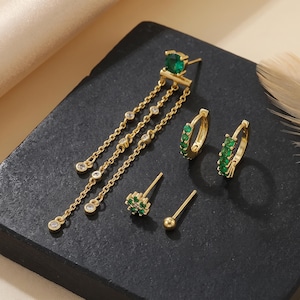 May include: A set of gold-tone earrings including one long dangle earring with a green stone and three chains with clear stones, two small hoop earrings with green stones, one flower-shaped stud with green stones, and one simple ball stud.