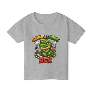 以下が含まれることがあります： 「Grumpasaurus Rex」の文字と、不機嫌そうな緑色の恐竜のイラストが描かれたグレーのTシャツ。火山、雲、先史時代の風景がデザインされています。柔らかい素材で作られたTシャツです。