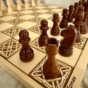May include: A wooden chess set with light and dark brown pieces on a light-colored board. The board features a Celtic knot design and the name "Jones" is engraved on the side. The chess pieces are arranged in the starting positions.