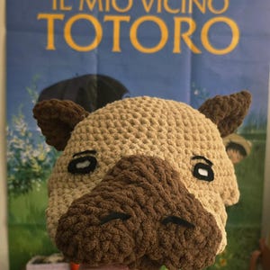 May include: A crocheted Totoro hat in beige and brown, featuring large ears and a textured snout. The hat has black eyes and is inspired by the animated film "My Neighbor Totoro". The background includes a movie poster.