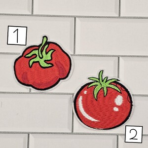 May include: Two embroidered tomato patches. One tomato patch is a rounded shape, and the other is a more oblong shape. Both patches are red with green stems and leaves. The patches are outlined in black and have a white border.