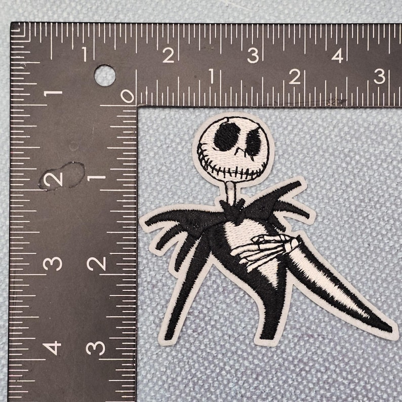 May include: Embroidered patch of Jack Skellington from The Nightmare Before Christmas. The patch is roughly 7.6 cm tall and 5 cm wide. The design is black and white, depicting a detailed skeleton figure.
