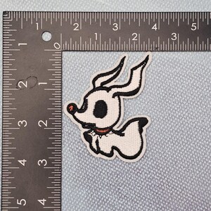 May include: An embroidered patch of a cartoon dog in white and black, with a red collar and a black eye. The patch measures approximately 5 cm in height and 5 cm in width.