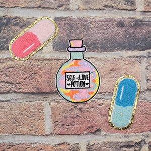 May include: Three embroidered patches on a brick background. One is a pink and white pill shape with gold trim. Another is a potion bottle with the text "SELF LOVE POTION". The third is a blue and white pill shape with gold trim.