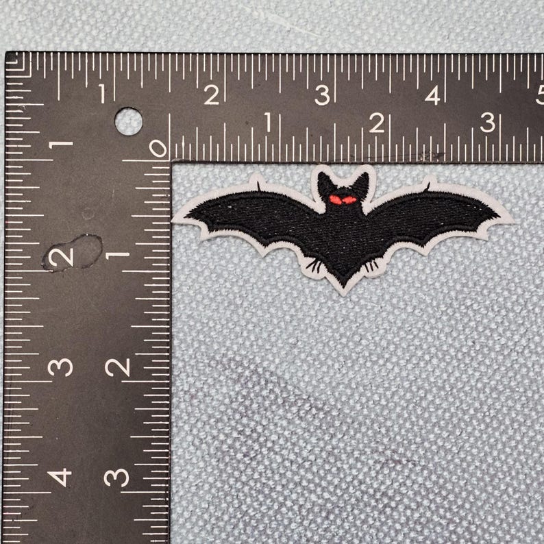 May include: Embroidered black bat patch with red eyes and a white border. The bat's wings are spread wide. The patch is approximately 7.6 cm wide. Perfect for Halloween or gothic-themed crafts, this patch can be ironed on or sewn onto fabric.