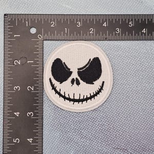 May include: A circular, white patch with a black, stylized face, including large eyes and a stitched grin. The patch is approximately 5 cm in diameter, set against a light blue background.