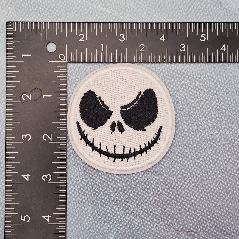 May include: A circular, white patch with a black, stylized face, including large eyes and a stitched grin. The patch is approximately 5 cm in diameter, set against a light blue background.