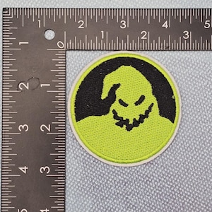 May include: Circular embroidered patch with a black character face on a lime green base. The patch has a white border and is approximately 7.6 cm in diameter. The design is a cartoonish representation of a face.