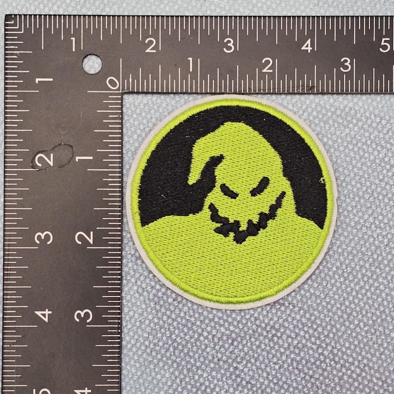 May include: Circular embroidered patch with a black character face on a lime green base. The patch has a white border and is approximately 7.6 cm in diameter. The design is a cartoonish representation of a face.