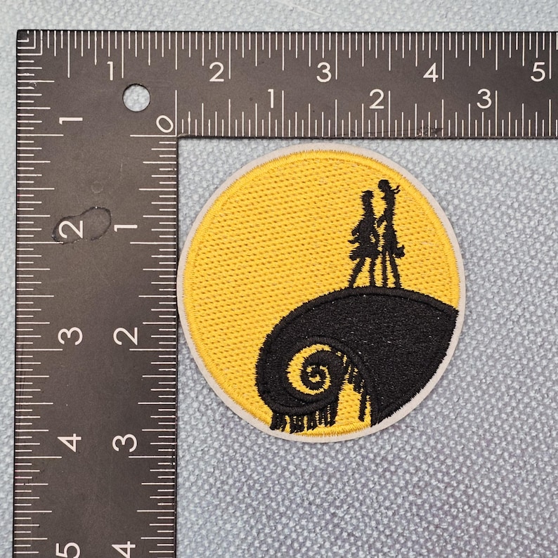 May include: Circular embroidered patch with a black silhouette of two figures on a hill against a yellow backdrop, and a black swirl design. The patch is approx. 7.6 cm in diameter and has a white border.