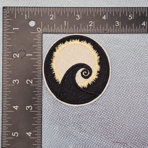 May include: Circular embroidered patch with a black and cream design. The patch shows a swirling black shape against a cream background, encircled by a cream border. The patch is approximately 7.6 cm in diameter.
