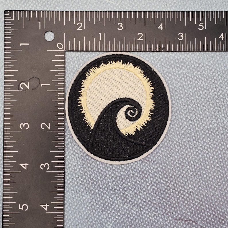May include: Circular embroidered patch with a black and cream design. The patch shows a swirling black shape against a cream background, encircled by a cream border. The patch is approximately 7.6 cm in diameter.