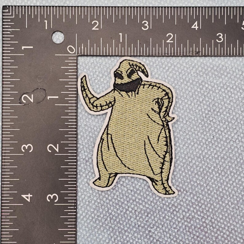 May include: An embroidered patch featuring a character with a stitched texture, in shades of tan and black. The character has a rounded body, a stitched face, and is posed with one arm raised. The patch is approximately 7.6 cm tall.