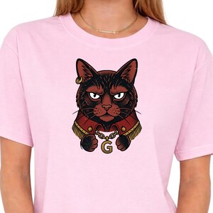 May include: A light pink t-shirt featuring a detailed illustration of a black cat with a stern expression. The cat wears a red jacket with gold trim and a gold chain with the letter "G".