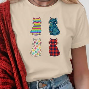 Könnte beinhalten: Cremefarbenes T-Shirt mit vier Cartoon-Katzen. Jede Katze hat ein einzigartiges Design: Regenbogenstreifen, Paisley, Tupfen und rot-schwarzes Karomuster. Die Katzen sitzen aufrecht.