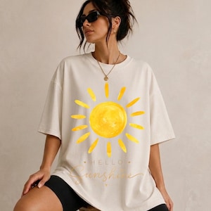 Può includere: T-shirt color crema con un grande disegno di sole giallo ad acquerello. La maglietta ha le parole "Hello Sunshine" in un carattere corsivo. La maglietta ha una vestibilità casual e oversize.