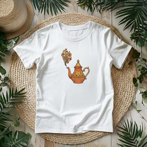 Puede incluir: Camiseta blanca con una colorida ilustración de una tetera. La tetera es naranja y amarilla con patrones decorativos. Un remolino de vapor verde, naranja y amarillo se eleva de la boquilla. La camiseta se muestra sobre una estera tejida.