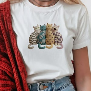 Könnte beinhalten: Weißes T-Shirt mit einer Grafik von vier stilisierten Katzen mit einzigartigen Mustern und Farben. Die Katzen sind eng beieinander angeordnet. Das Shirt wird mit einer blauen Jeans und einem roten Cardigan getragen.